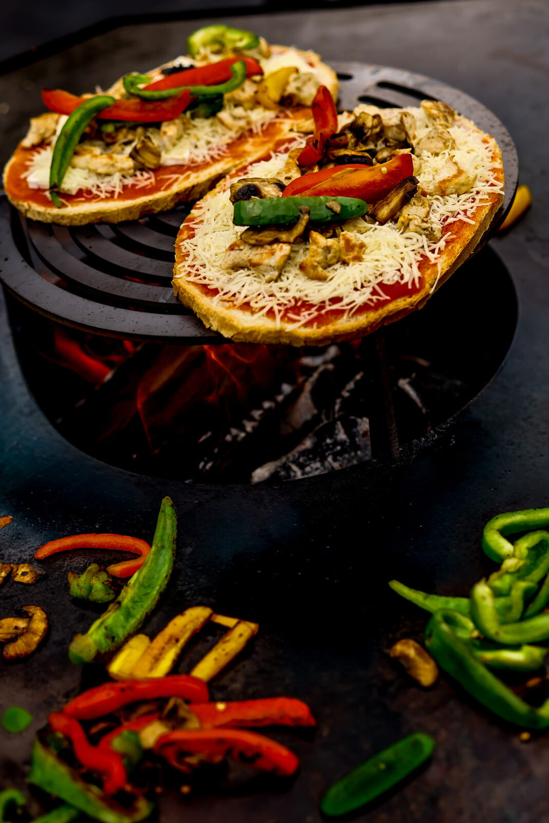 Pizzas garnies posées sur la grille du foyer, braises visibles en dessous