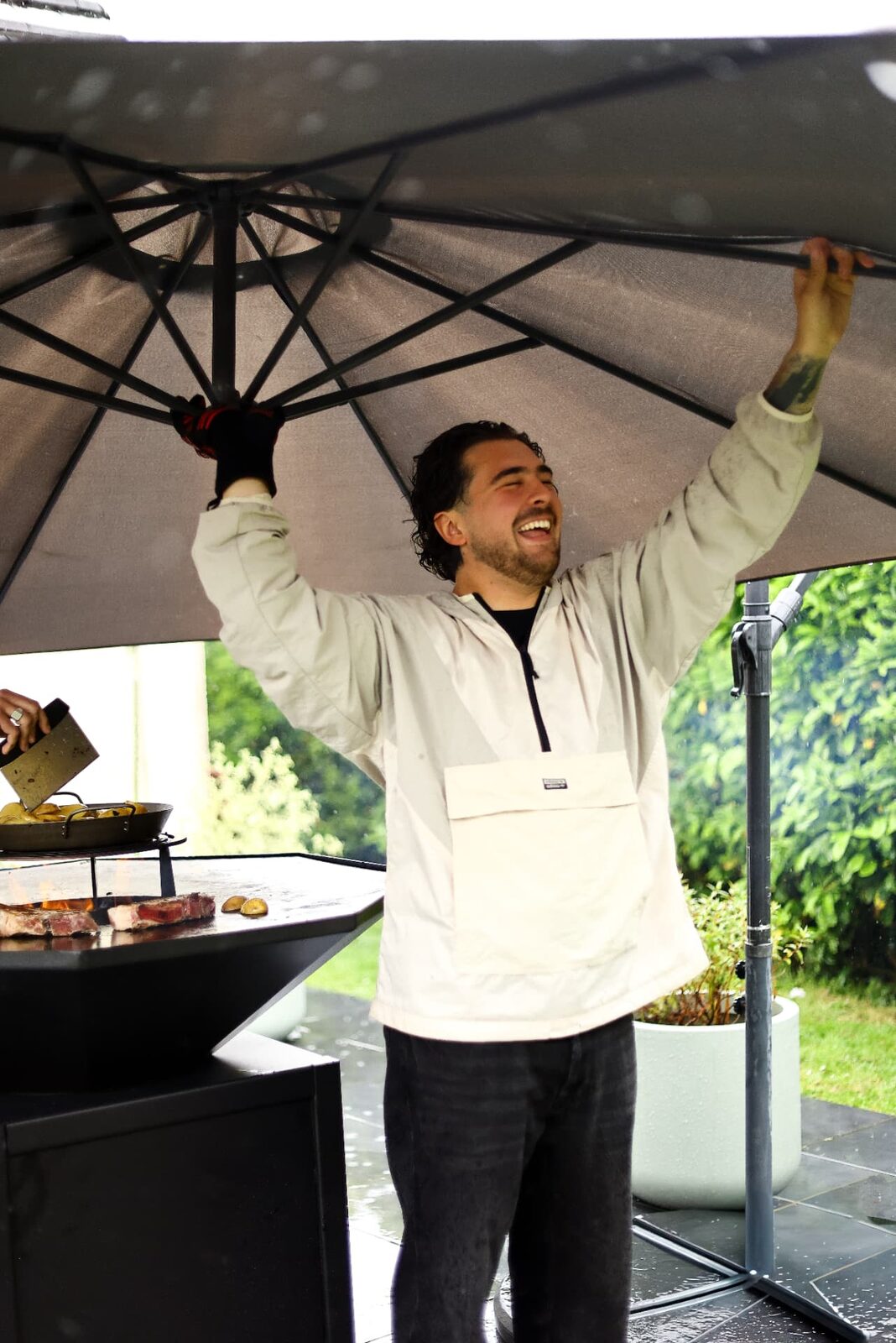 Le cuisinier, sourire aux lèvres, ajuste le parasol au-dessus du brasero sous la pluie